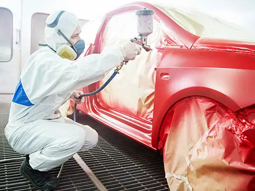 A worker dressed in protective gear sprays a coat of red paint on the exterior of an automobile.